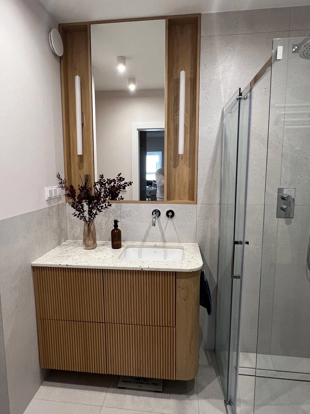Waschtischunterschrank nach Maß mit gerillten Eiche-Fronten und abgerundeter Ecke, darauf eine Terrazzo-Platte mit Unterbaubecken; darüber ein Spiegel im Eichenrahmen zwischen zwei schmalen LED-Leuchten, rechts die Dusche.
