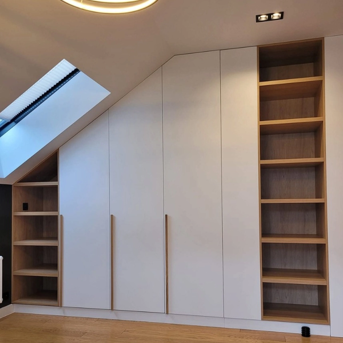 Einbauschrank unter einer Dachschräge: grifflose weiße Türen folgen der Dachneigung, geöffnet über schmale Eichenleisten an den Kanten, an beiden Enden offene Regalfächer in Eiche — links niedrig unter dem Dachfenster, rechts als hohe Regalsäule.