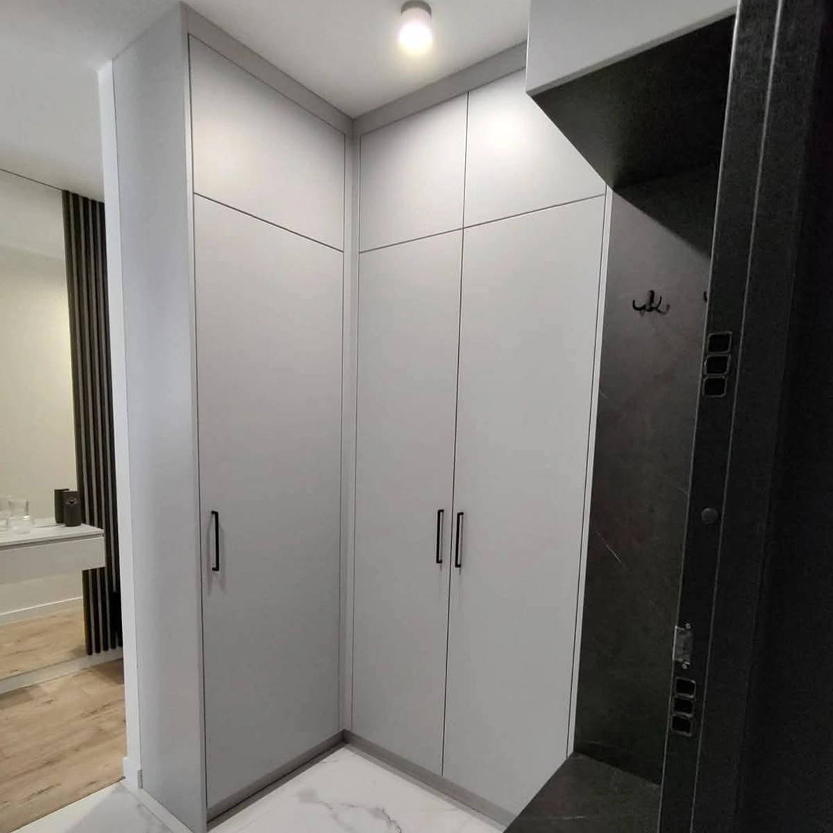 Deckenhoher Einbauschrank in einer Flurecke: matte hellgraue Fronten mit schwarzen Griffleisten führen um die Ecke herum, rechts daneben eine dunkle Wandverkleidung mit Garderobenhaken.