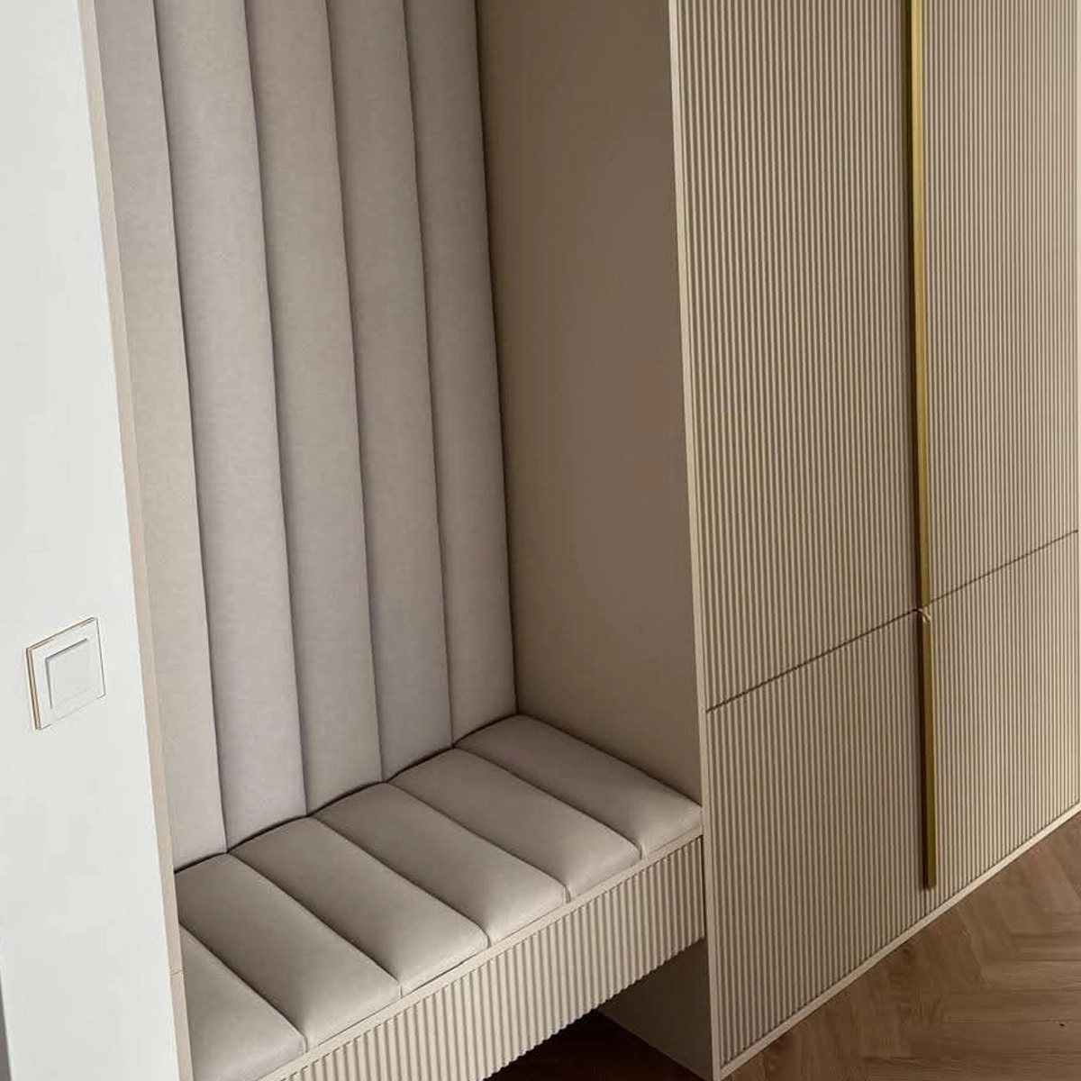 Detail einer Garderobe nach Maß: offene Sitznische mit gepolsterten Wandpaneelen und passendem Sitzpolster, daneben die gerillte Schrankfront mit schmaler goldener Griffleiste.