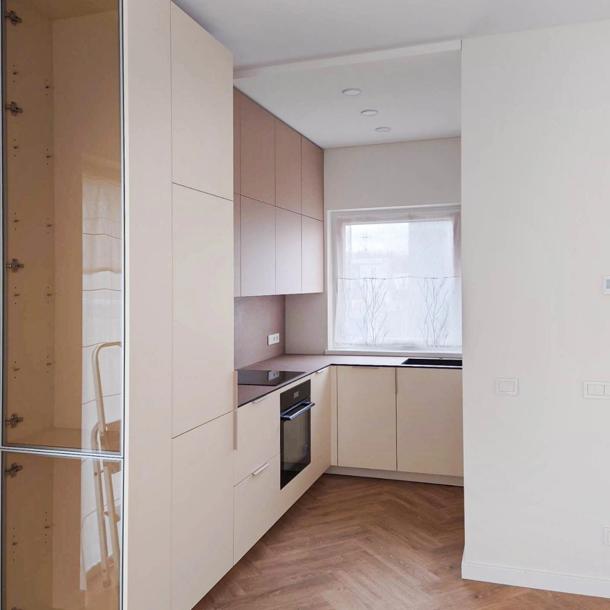 Kompakte Eckküche nach Maß in Beige: grifflose Fronten bis unter die Decke, Hochschrank mit Einbaubackofen und eine Arbeitsplatte, die unter dem Fenster durchläuft.