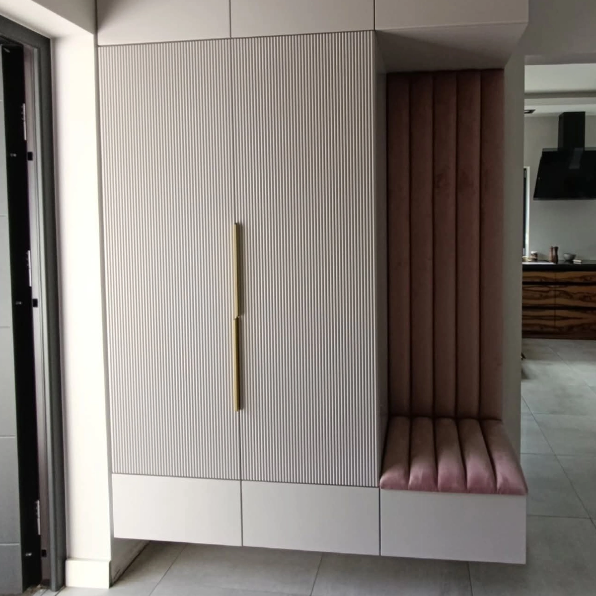 Schwebender Garderobenschrank im Flur mit gerillten hellen Fronten und langem goldenem Griff, rechts daneben eine Sitzbank mit gepolstertem Rückenpaneel in Altrosa.